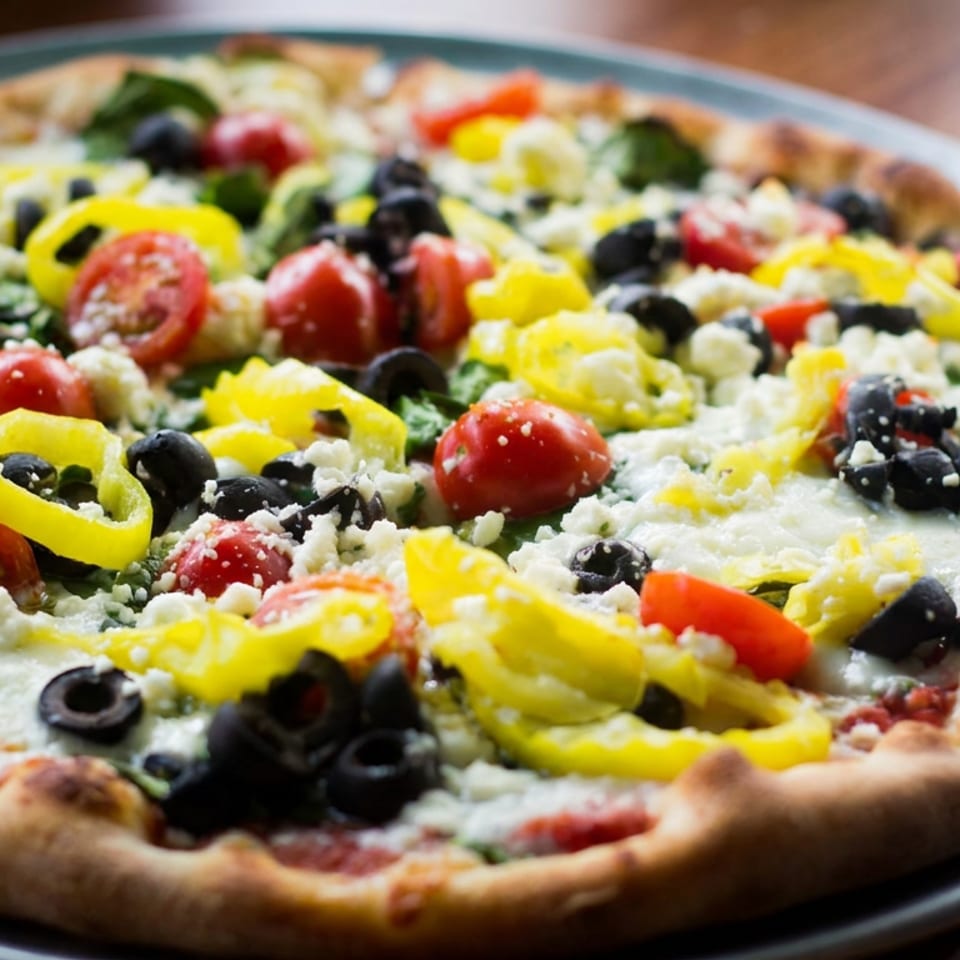 Close-up of a loaded vegetable pizza topped with tomatoes, yellow peppers, black olives, greens, and crumbled cheese.