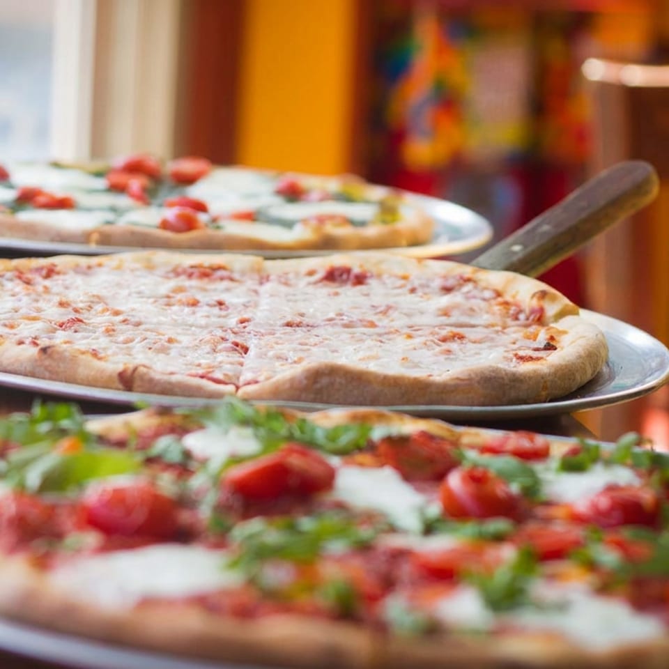 Three pizzas on metal serving trays, including cheese pizza and pizzas topped with tomatoes, herbs, and melted cheese.