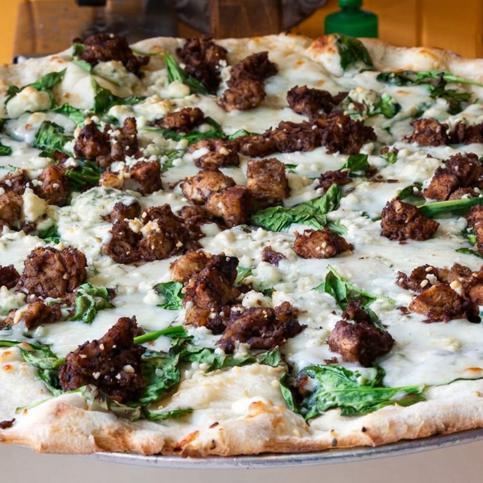 Thin-crust pizza topped with creamy cheese, leafy greens, and browned chunks of meat on a metal serving tray.
