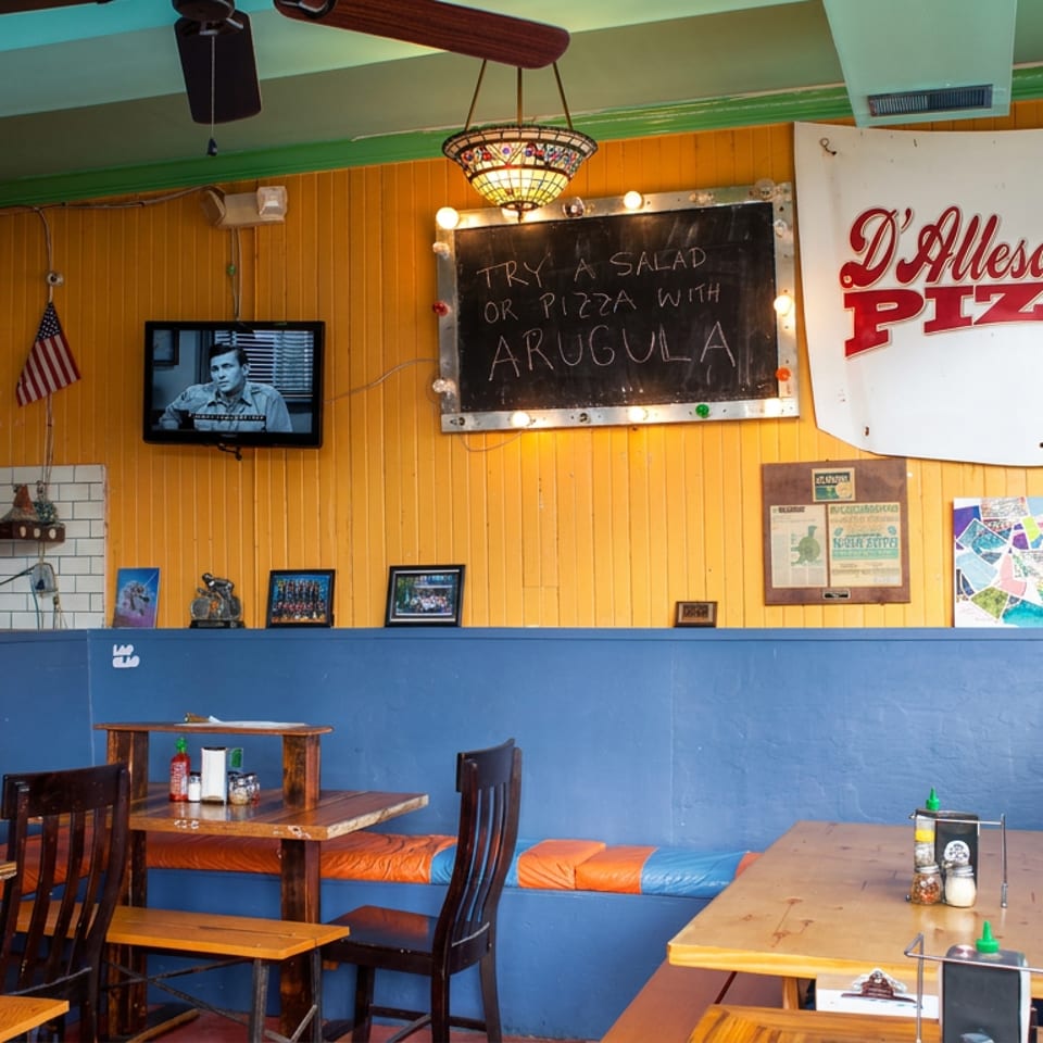 Colorful pizzeria interior with blue booths, wooden tables, wall art, a hanging lamp, bicycle, television, and D’Alessandro Pizza sign.