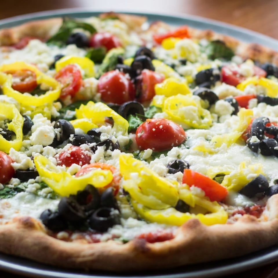 Vegetable pizza topped with cherry tomatoes, black olives, banana peppers, spinach, and crumbled cheese on a metal tray.