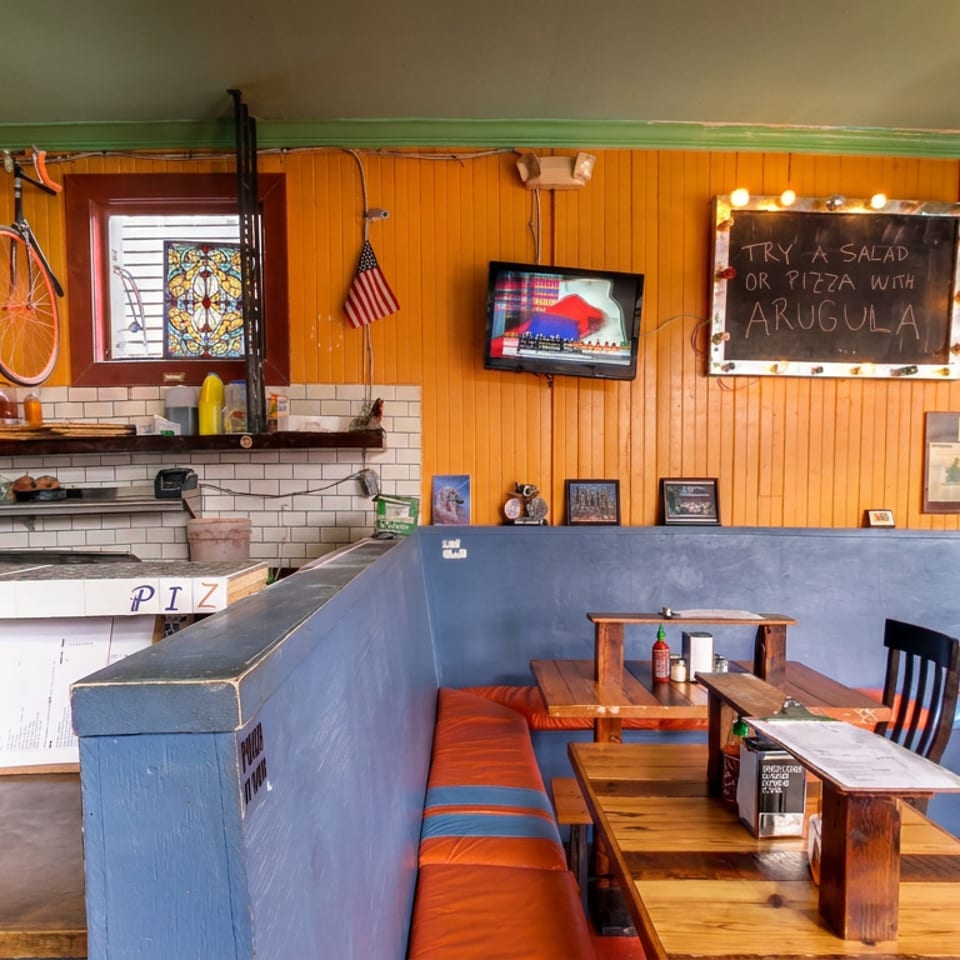 Colorful pizzeria interior with blue booths, wooden tables, orange walls, pizza counter, bicycle decor, and chalkboard menu.