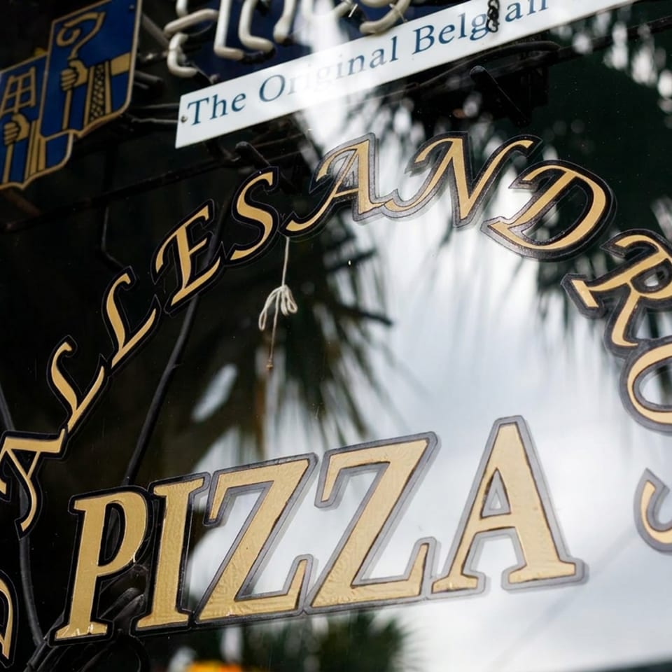 Restaurant window displays ornate lettering reading “Jasand Pizze” and “The Original Belgian,” reflecting palm trees.