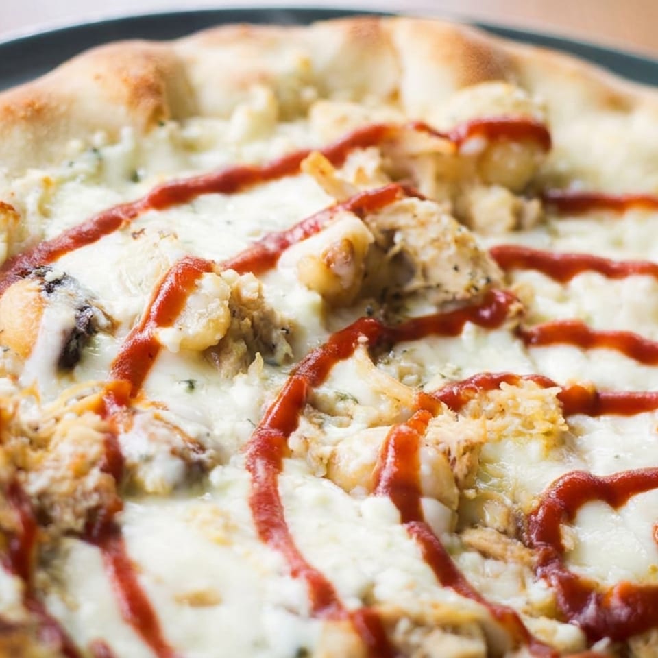 Close-up of a cheesy pizza topped with chicken, mushrooms, and red sauce drizzled across the surface.