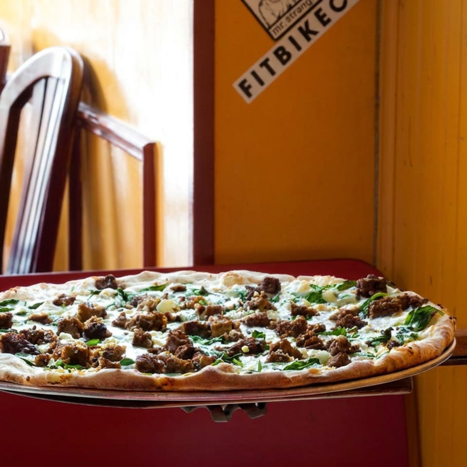 Freshly baked thin-crust pizza topped with cheese, greens, and browned meat sits on a wooden pizza peel in a cozy restaurant.